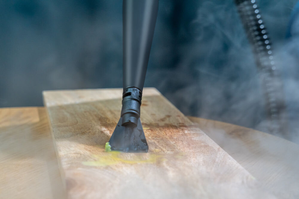 ODKURZACZ PIORĄCY SPOTCLEAN GOTZE&JENSEN CSC600 ze szczotką uniwersalną szeroką do butów do czyszczenia tapicerki meblowej samochodowej dywanów Zbliżenie na małą, czarną ssawkę parową emitującą parę i czyszczącą jaskrawożółtą plamę na drewnianej powierzchni (prawdopodobnie blacie).