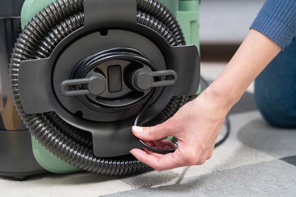 ODKURZACZ PIORĄCY SPOTCLEAN GOTZE&JENSEN CSC600 z funkcją prania osuszania usuwania nadmiaru wody szybkiego schnięcia tapicerki Zbliżenie na rękę kobiety, która zwija czarny kabel zasilający na wbudowanym uchwycie z tyłu odkurzacza. Widać również zwinięty wąż elastyczny.