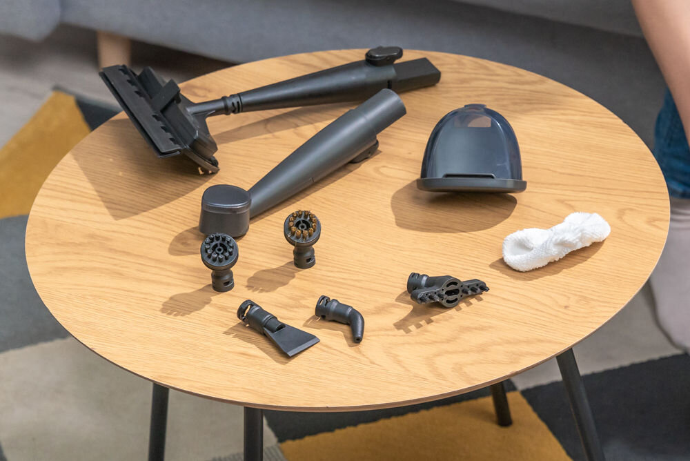 ODKURZACZ PIORĄCY SPOTCLEAN GOTZE&JENSEN CSC600 Zestaw akcesoriów do odkurzacza piorącego lub parownicy, rozłożony na drewnianym, okrągłym stoliku kawowym. Akcesoria są w kolorze czarnym/antracytowym i obejmują dużą nasadkę do podłóg, różne szczotki i dysze do detali, małą końcówkę i małą, białą ściereczkę z mikrofibry.