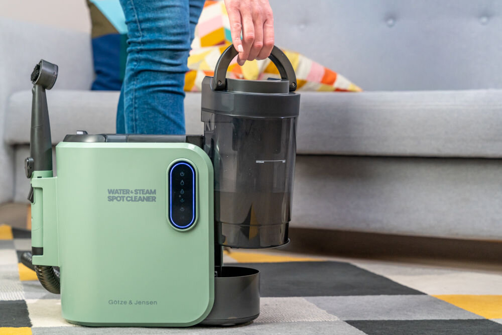 ODKURZACZ PIORĄCY SPOTCLEAN GOTZE&JENSEN CSC600 z funkcją para plus zasysanie 13 kPa do usuwania starych plam wina tłuszczu oleju z tapicerki dywanów Kobieta podnosi za uchwyt czarno-zielony odkurzacz piorący Götze & Jensen Water+Steam Spot Cleaner stojący na dywanie. Odkurzacz ma czarny pojemnik na wodę i panel sterowania z podświetlanymi ikonami.