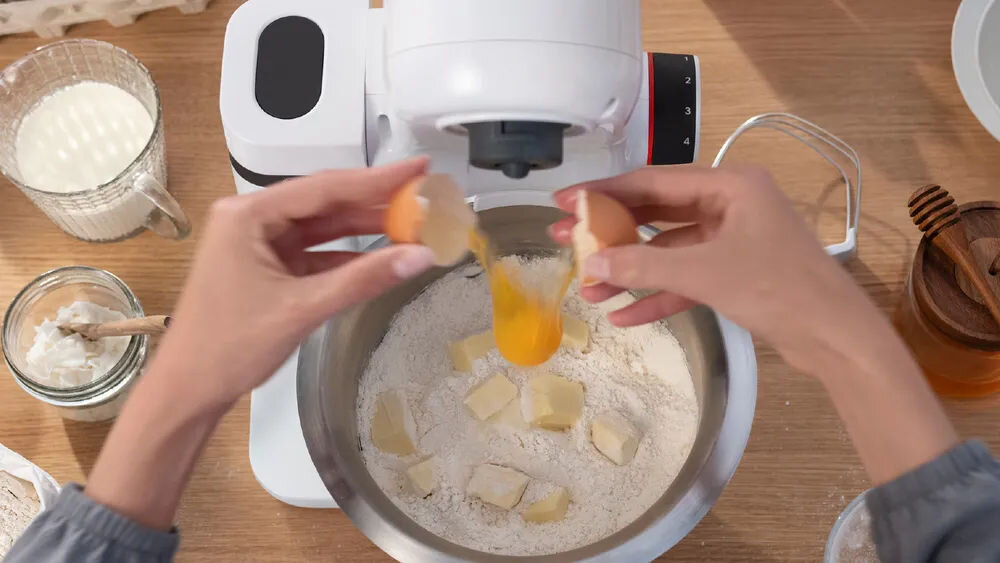 Robot kuchenny planetarny BOSCH MUMS2EW11 z blenderem kielichowym robot biała obudowa osoba wbija jajka do misy misa o pojemności 3,8 litra z Tritanu nie pochłania zapachów komfort użytkowania specjalnie zaprojektowany kształt dokładne wymieszanie składników wygoda funkcjonalność