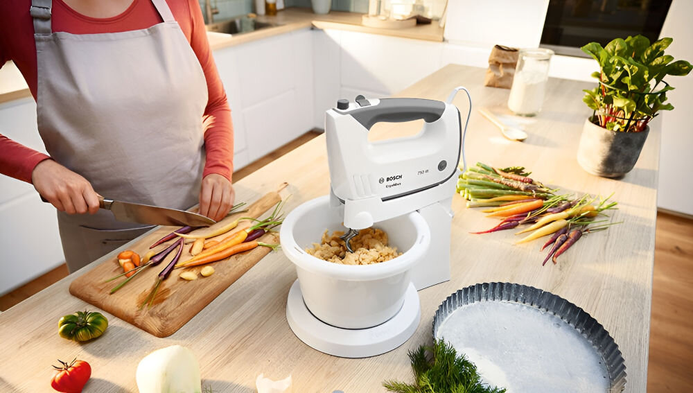 Mikser BOSCH ErgoMixx MFQ37490 Biały blender stoi na blacie misa biała obudowa miksowanie składników szare elementy osoba kroi warzywa na desce stabilny stojak misa o pojemności 1 litra obracająca się automatycznie mieszanie bez konieczności użycia rąk dokładne równomierne wymieszanie składników podczas przyrządzania mniejszych porcji ciasta