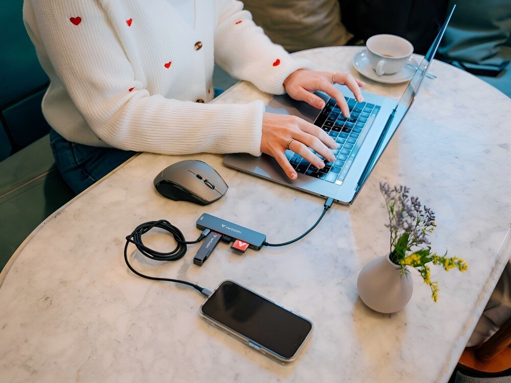 Hub Verbatim 32156 Widok z góry na osobę piszącą na laptopie. Do urządzenia podłączony jest hub USB-C z myszką, pendrive'em, czytnikiem kart i telefonem, 
            hub usba 3.2 gen1 5gb/s przesyłanie usbc power delivery pd ładowanie