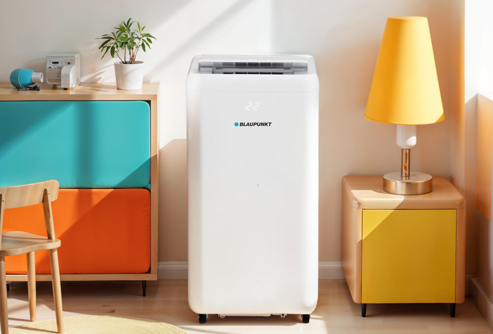 Biały klimatyzator przenośny z logo BLAUPUNKT stoi w jasnym pomieszczeniu obok kolorowej komody i żółtej lampy. Na wyświetlaczu klimatyzatora widoczna jest liczba \'22\'.
