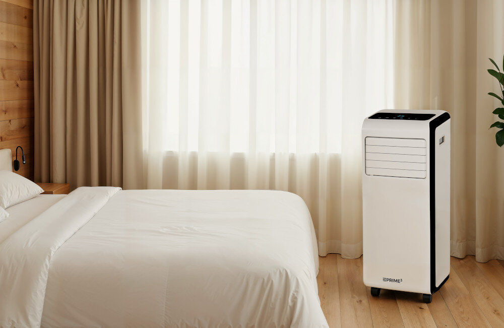 W jasnym pokoju z łóżkiem znajduje się biały klimatyzator przenośny na kółkach z widocznym logo \'PRIME3\'.