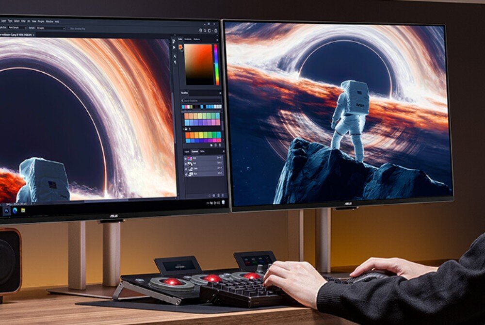 Na biurku znajdują się dwa monitory ASUS, na których wyświetlany jest obraz kosmonauty stojącego przed czarną dziurą. Przed monitorami widoczna jest klawiatura i panel sterujący, a osoba obsługuje komputer.