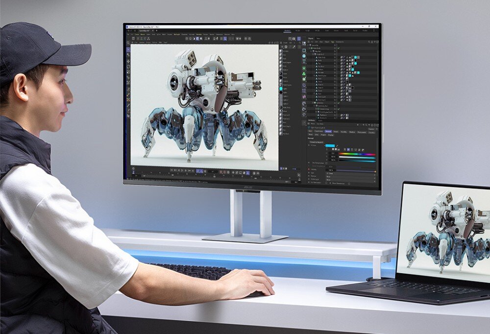 Osoba siedzi przy biurku, patrząc na duży monitor, na którym wyświetlany jest obraz robota w programie graficznym. Obok monitora znajduje się laptop z tym samym obrazem na ekranie.