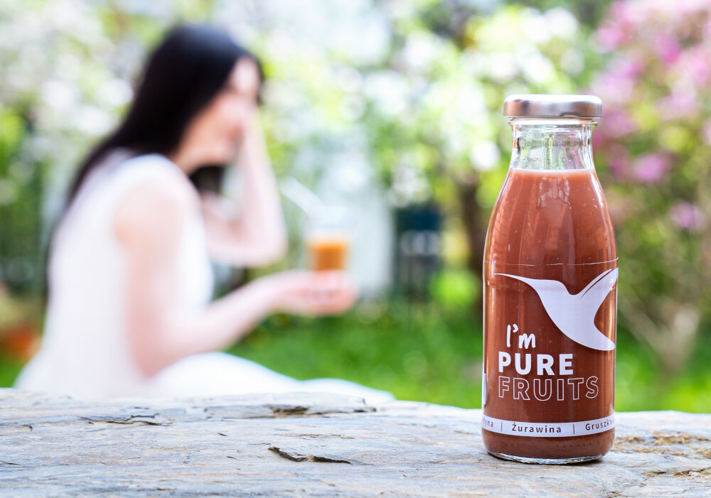 Sok COLLIBRE Pure Fruits Jeżynowo-gruszkowo-żurawinowy 15 x 250 ml butelka soku ustawiona na kamiennym podłożu w tle siedzi osoba kwiaty krzewy dom naturalny smaczny forma przecierowa orzeźwiający