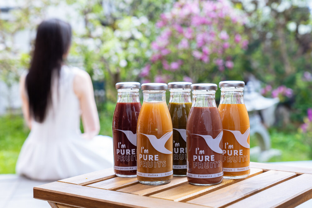 Sok COLLIBRE Pure Fruits Mix Smaków 15 x 250 ml butelki soku ustawione na drewnianym podłożu w tle siedzi osoba kwiaty krzewy dom naturalne soki smaczny forma przecierowa orzeźwiające zdrowie