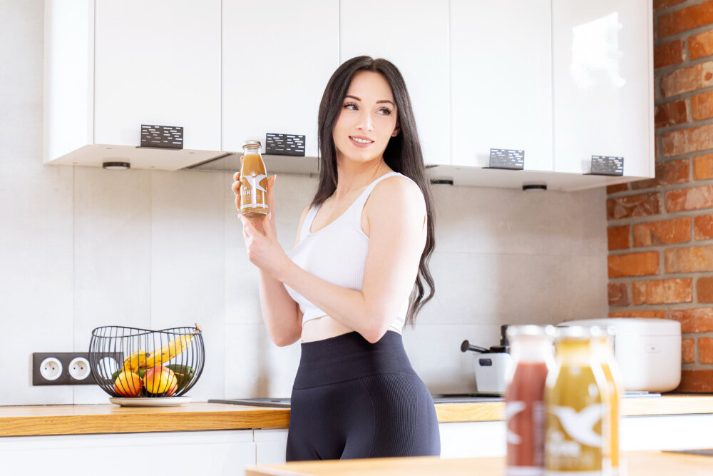 Sok COLLIBRE Pure Fruits Mix Smaków 15 x 250 ml kuchnia kobieta stoi przy blacie w dłoni trzyma butelkę poręczne butelki zmieszczą się w torbie czy plecaku pyszny zdrowy napój naturalna słodycz owoców dla dzieci i dorosłych soki NFC bezpieczny zdrowy wybór