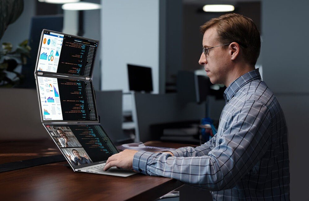 Monitor ASUS ZenScreen Duo MQ149CD Mężczyzna w okularach siedzi przy biurku z laptopem i monitorem z dwoma ekranami ustawionymi pionowo, na ekranach kod programistyczny, wykresy i wideokonferencja, widoczność, czas reakcji, redukcja migotania, filtr niebieskiego światła