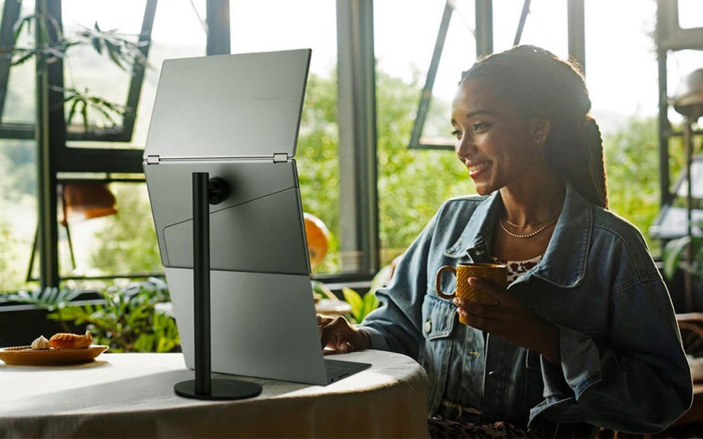 Monitor ASUS ZenScreen Duo MQ149CD Kobieta siedzi przy stole z filiżanką w dłoni, przed nią laptop i monitor z dwoma ekranami ustawiony na statywie, w tle duże okno i rośliny, obracanie, orientacja pionowa i pozioma, montaż na statywie