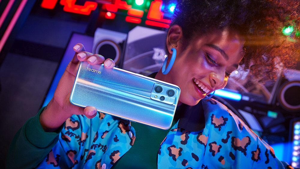 Osoba trzyma smartfon z logo \'realme\', w tle kolorowe światła i sprzęt audio.