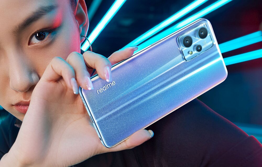 Osoba trzyma smartfon z widocznym logo \'realme\' na tylnej obudowie. Telefon ma błyszczące wykończenie i trzy kamery w module z tyłu.