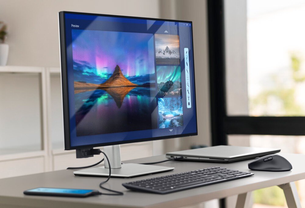 Monitor DELL UltraSharp U2725QE Stanowisko pracy w jasnym pomieszczeniu z monitorem, na którym otwarty jest program graficzny z miniaturami zdjęć zorzy polarnej, obok stoi otwarty laptop, klawiatura, mysz i smartfon na pierwszym planie, przekątna, rozdzielczość, matryca
