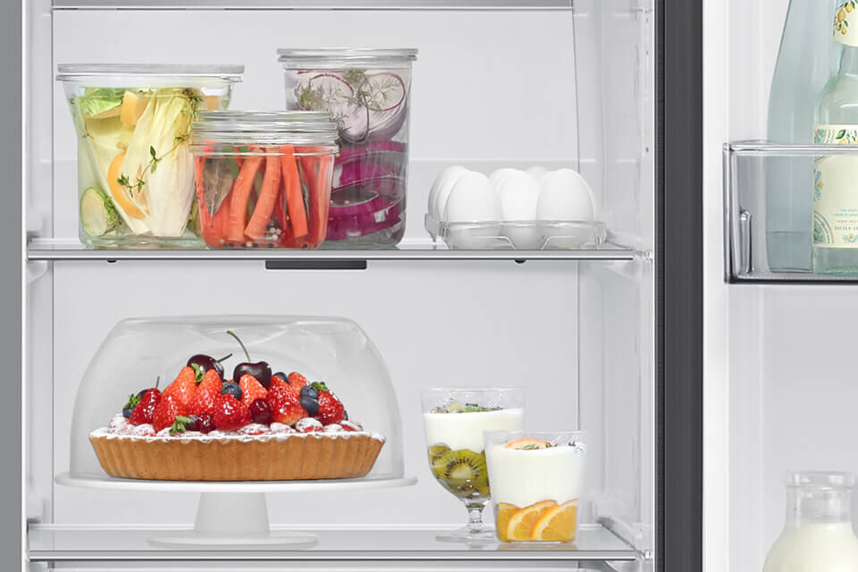 rs80f66kbfeo półki z hartowanego szkła Wnętrze lodówki z półkami, na których znajdują się różne produkty spożywcze, w tym ciasto z owocami, słoiki z warzywami, jajka oraz jogurty z owocami. Na drzwiach widoczna jest butelka z napojem.