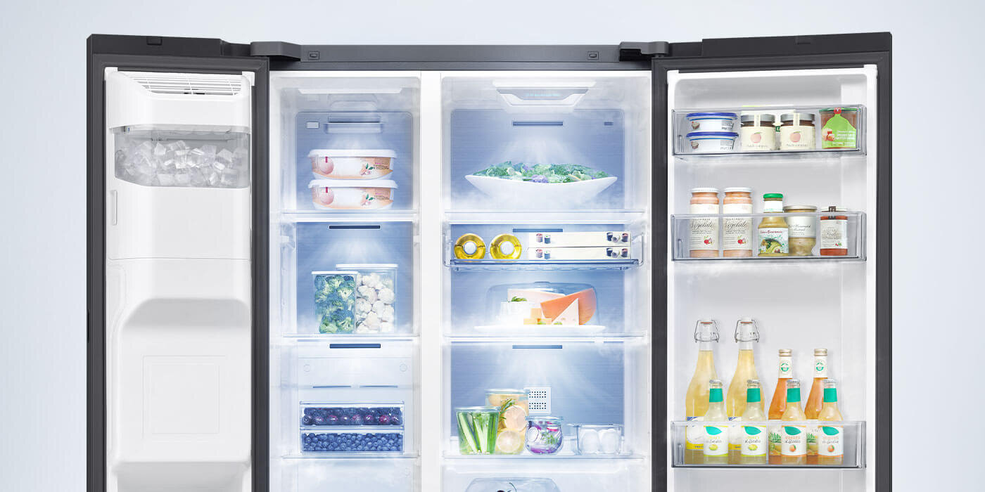 Otwarta lodówka z widocznymi półkami wypełnionymi różnymi produktami spożywczymi, takimi jak napoje, warzywa i słoiki. Po lewej stronie znajduje się kostkarka do lodu.