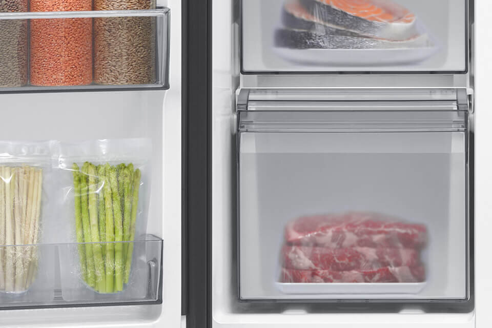 Wnętrze lodówki z widocznymi półkami, na których znajdują się produkty spożywcze, takie jak słoiki z kaszami, zapakowane szparagi oraz mięso w szufladzie. Obok znajduje się zamrażarka z zamrożonymi rybami i mięsem.