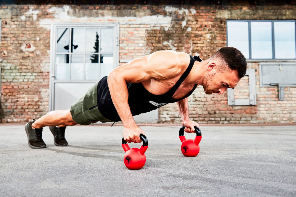 Kettlebell GORILLA SPORTS 3457 (10 kg) sala treningowa siłownia osoba w stroju sportowym robi pompki opiera dłonie na kettlebellach w tle ściana z cegły siła kontrola do codziennego użytku zdrowie