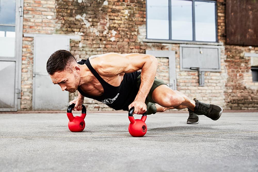 Kettlebell GORILLA SPORTS 3457 (10 kg) sala treningowa siłownia osoba w stroju sportowym robi pompki opiera dłonie na kettlebellach w tle ściana z cegły ćwiczenia wymagają utrzymania balansu i precyzyjnej kontroli ruchu nieregularny rozkład ciężaru zmusza do aktywnej pracy mięśni poprawić postawę płynność ruchów