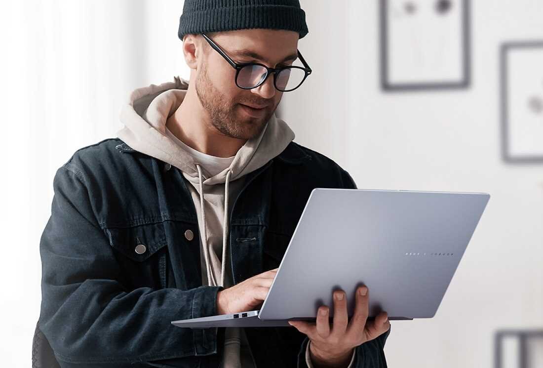 Mężczyzna w ciemnej czapce, okularach i jeansowej kurtce z beżową bluzą z kapturem trzyma w dłoniach srebrnego laptopa ASUS VivoBook S 14, koncentrując się na ekranie; w tle jasne wnętrze z białymi ścianami i oprawionymi obrazami, naturalne światło z okna. - cechy zalety opis przeznaczenie