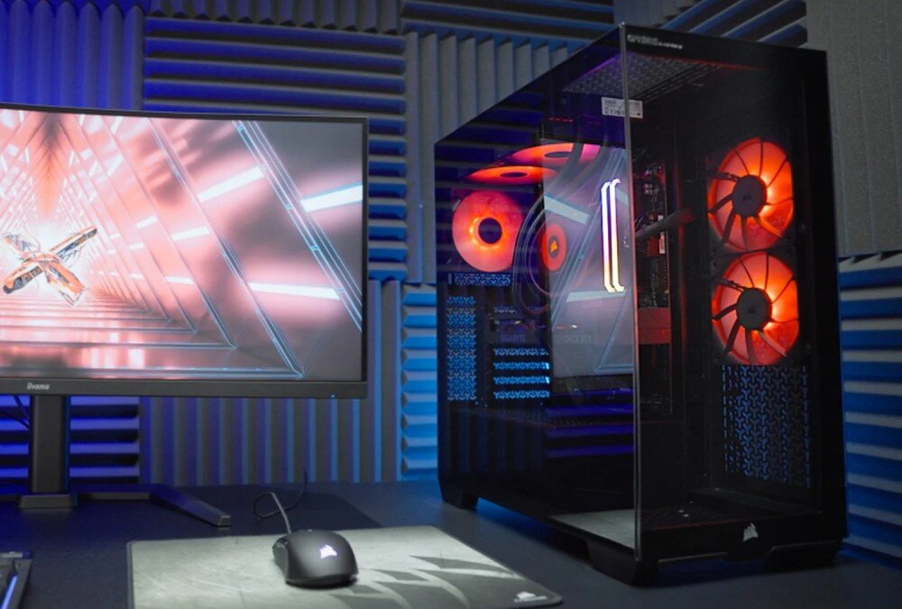 Komputer OPTIMUS E-sport GA550T-CR5 Komputer stacjonarny z przezroczystym panelem bocznym i czerwonym podświetleniem wnętrza stoi na biurku obok monitora, na którym trwa rozgrywka w grę FPS. W tle widać panele akustyczne na ścianie, opis