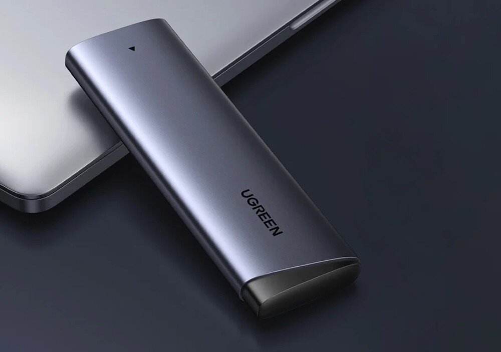 Obudowa dysku UGREEN CM767 Podłużna, metalowa obudowa na dysk w kolorze szarym, ustawiona pionowo pod skosem na ciemnym tle. Na obudowie widoczne jest logo Ugreen, port USB-C na górze oraz mała dioda kontrolna