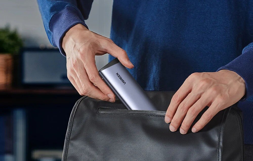 Obudowa dysku UGREEN CM767 Mężczyzna w niebieskiej bluzie wkłada dłonią szarą obudowę do małej kieszeni czarnej torby podróżnej, co ilustruje kompaktowy rozmiar urządzenia