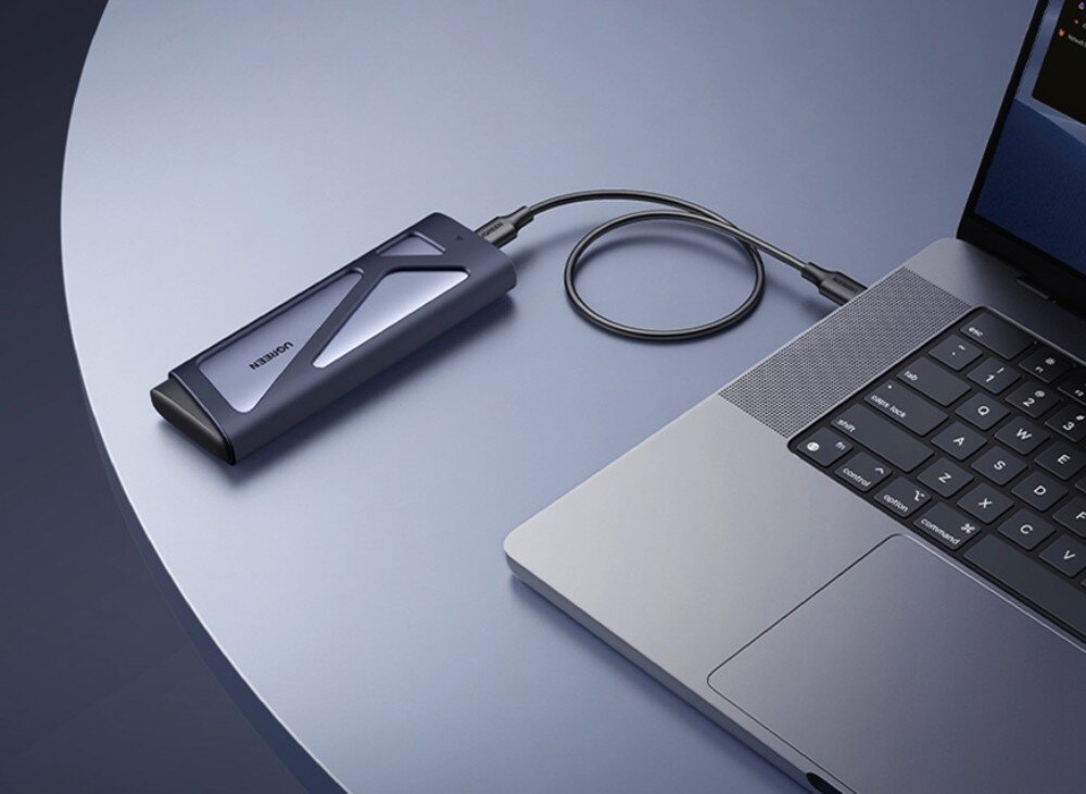 Obudowa dysku UGREEN CM767 Podłużna, metalowa obudowa na dysk w kolorze szarym, ustawiona pionowo pod skosem na ciemnym tle. Na obudowie widoczne jest logo Ugreen, port USB-C na górze oraz mała dioda kontrolna