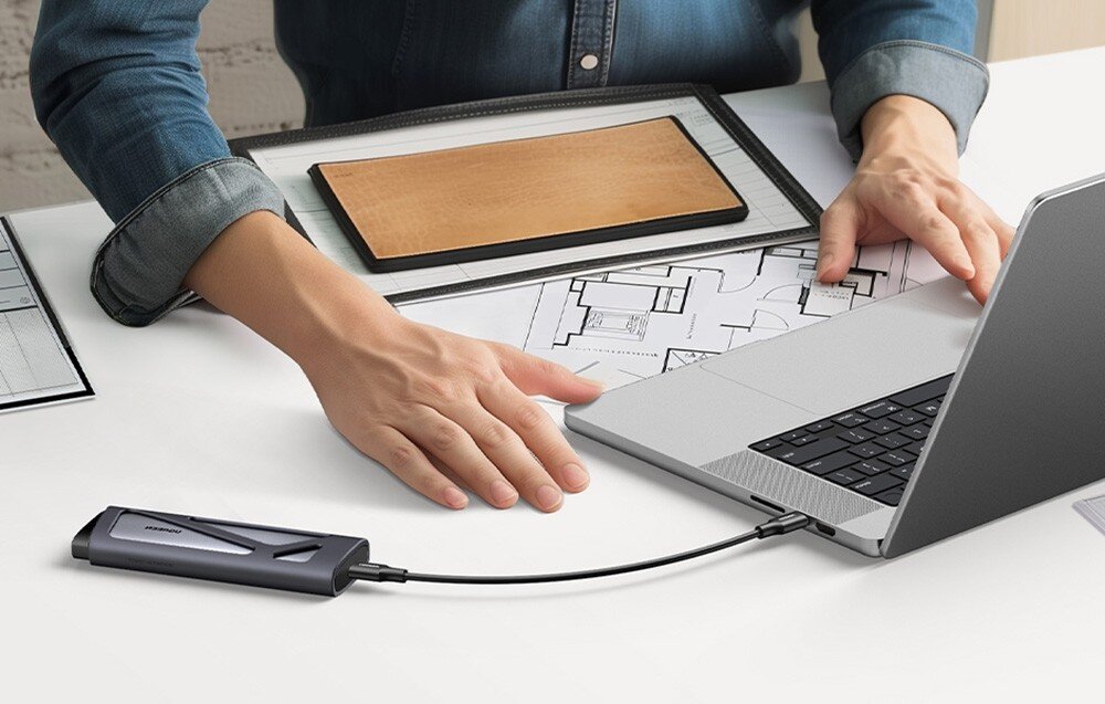 Obudowa dysku UGREEN CM767 Zbliżenie na szarą, matową obudowę spoczywającą na obudowie laptopa. Urządzenie ma zaokrąglone krawędzie, widoczne logo producenta oraz czarną zatyczkę na dolnym końcu