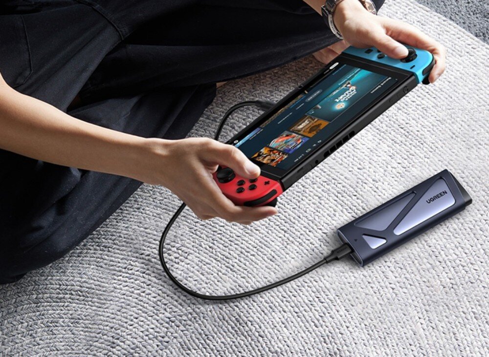 Obudowa dysku UGREEN CM767 Mężczyzna w niebieskiej bluzie wkłada dłonią szarą obudowę do małej kieszeni czarnej torby podróżnej, co ilustruje kompaktowy rozmiar urządzenia