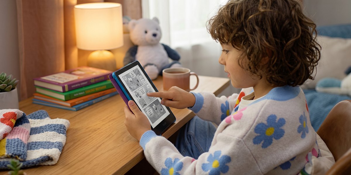 Dziecko siedzi przy biurku i używa czytnika e-booków, na którym wyświetlane są czarno-białe ilustracje. W tle znajduje się lampka, pluszowy miś i stos książek.
