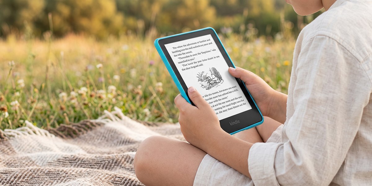 Osoba siedzi na kocu na trawie, trzymając czytnik e-booków z niebieską ramką. Na ekranie widoczny jest tekst i ilustracja.