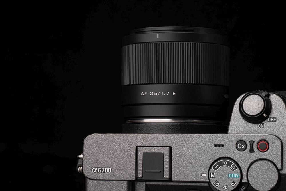Na zdjęciu widoczny jest aparat fotograficzny z zamontowanym obiektywem oznaczonym jako \'AF 25/1.7 E\'. Aparat ma oznaczenie \'α6700\' i widoczne są przyciski oraz pokrętła sterujące.