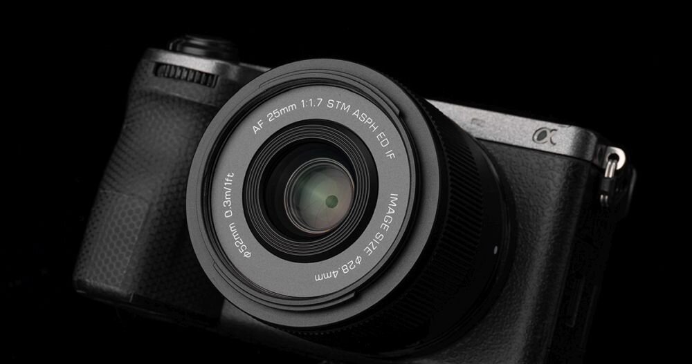 Na zdjęciu widoczny jest aparat fotograficzny z zamontowanym obiektywem. Na obiektywie znajduje się tekst: \'AF 25mm 1:1.7 STM ASPH ED IF\'.