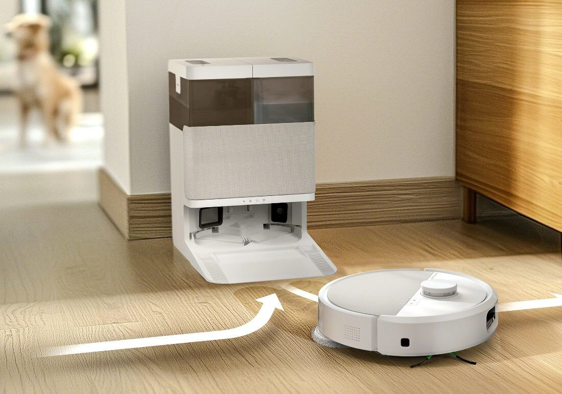 Na drewnianej podłodze znajduje się biały robot sprzątający obok stacji dokującej. W tle widoczny jest rozmyty obraz psa.