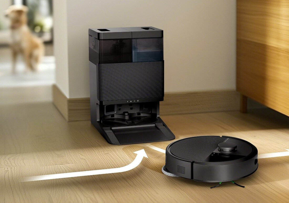 Na drewnianej podłodze znajduje się czarny robot sprzątający obok stacji dokującej. Na podłodze widoczna jest biała strzałka wskazująca kierunek ruchu robota.