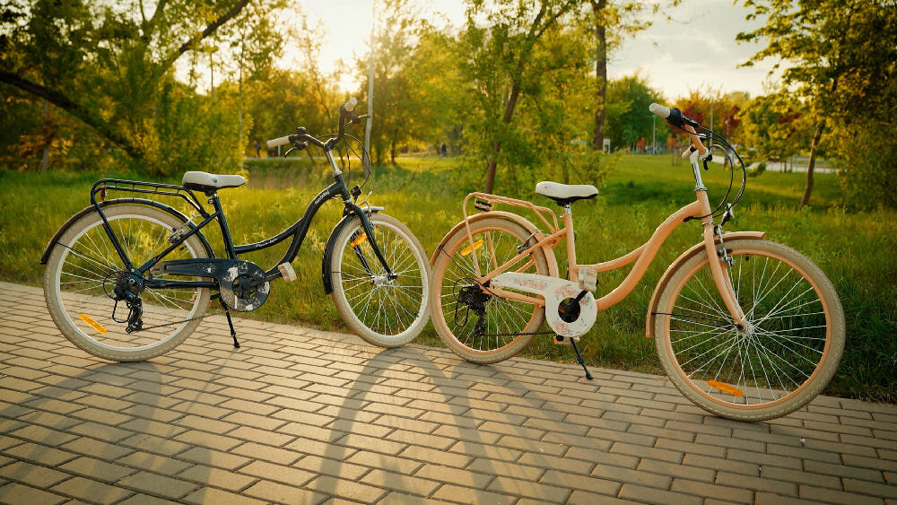 Rower młodzieżowy SUN BABY Flower Bike 24 cale dla dziewczynki Morelowy chodnik dwa rowery oparte o stopki w tle drzewa trawa stopka w centralnym punkcie ciężkości reflektory odblaskowe z przodu z tyłu i na szprychach roweru lepsza widoczność na drodze bezpieczeństwo dzwonek