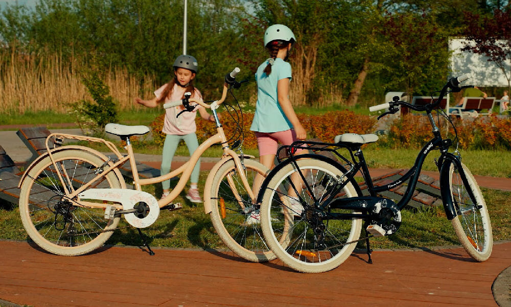 Rower młodzieżowy SUN BABY Flower Bike 24 cale dla dziewczynki Morelowy chodnik w parku dwa rowery oparte o stopki za rowerami dwie dziewczynki w tle drzewa 7 biegów obrotowa manetka Revoshift tylna przerzutka SHIMANO Tourney przerzutki SHIMANO duża wytrzymałość sprawne działanie płynna bezproblemowa zmiana biegów