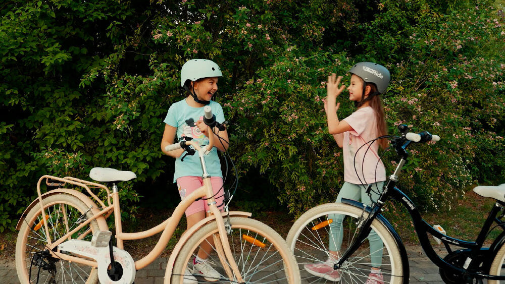 Rower młodzieżowy SUN BABY Flower Bike 24 cale dla dziewczynki Morelowy na pierwszym planie dwa rowery w tle dwie dziewczynki śmieją się do siebie drzewa wysoki komfort jazdy brak uczucia dyskomfortu odpowiednia pozycja ciała uchwyty z antypoślizgowego materiału regulacja w zakresie od 95 do 98 cm