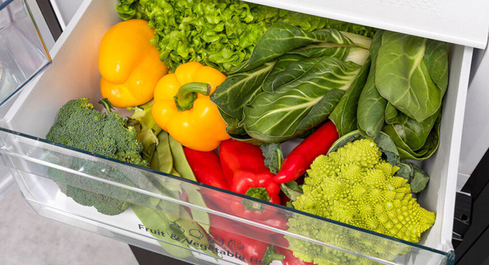Szuflada lodówki wypełniona różnymi warzywami, takimi jak papryka, sałata, brokuły i inne zielone warzywa. Na szufladzie widoczny jest napis \'Fruit & Vegetable Box\'.