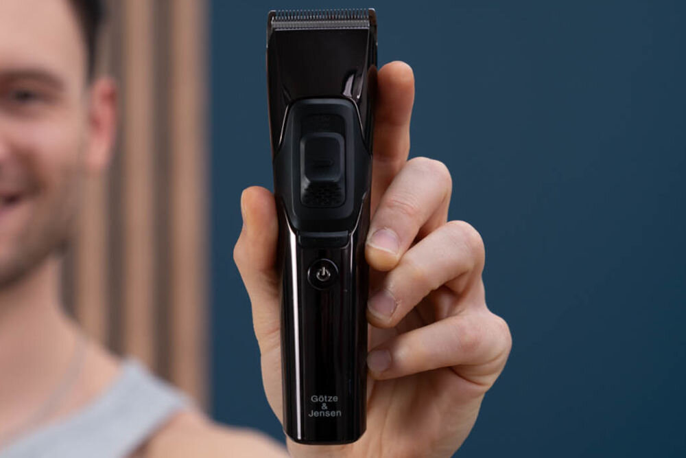 Strzyżarka GÖTZE & JENSEN CL950 0.8-2 mm Mężczyzna trzymający w dłoni czarną, błyszczącą maszynkę do strzyżenia włosów Götze & Jensen z widocznym przyciskiem zasilania i ostrzem tnącym.