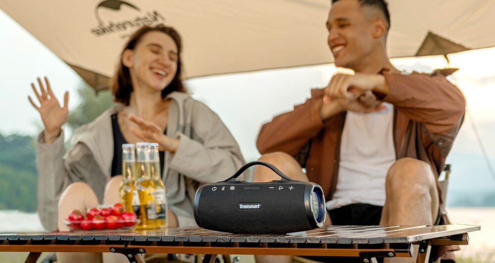 Głośnik TRONSMART Mirtune S100 moc 50W basy i czyste tony, głośnik stojący na stoliku, osoby siedzące na krzesłach w plenerze