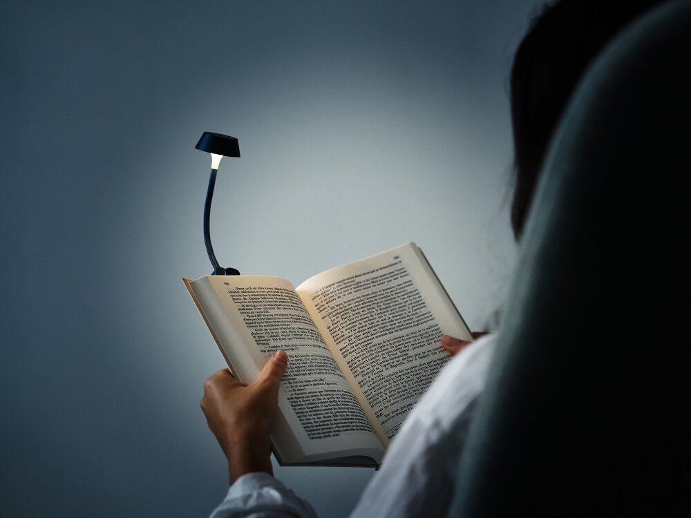 Lampka LED LEXON Mimi Ciemnoniebieski osoba trzyma książkę wpięta granatowa lampka do codziennego oświetlenia stylowy design praktyczność elastyczne ramię klips na książce biurku półce do domu biura w podróży dobre światło