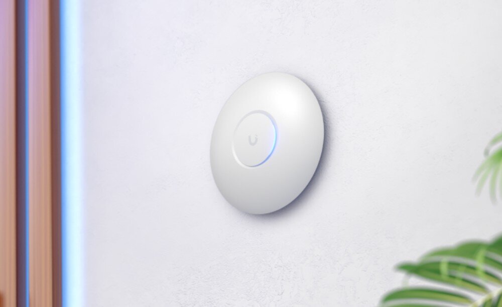 Punkt dostępu UBIQUITI U7-Lite Urządzenie sieciowe w kształcie białego dysku zawieszone na jasnej ścianie obok zielonych liści rośliny