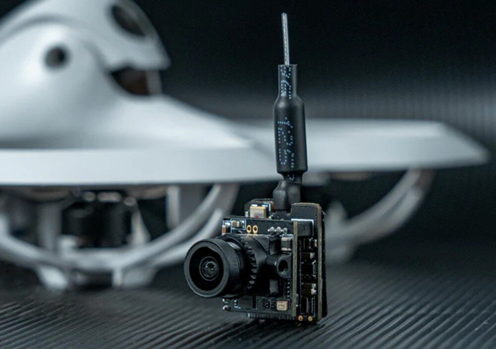 Kamera BETAFPV C04 + Moduł M04 VTX Czarny moduł kamery z anteną ustawiony pionowo na tle rozmytego białego korpusu drona