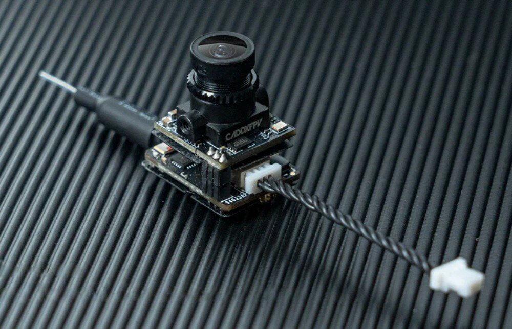 Kamera BETAFPV C04 + Moduł M04 VTX Moduł kamery z widocznym logo 'CADDXFPV' i podłączonym skręconym przewodem leżący na czarnej prążkowanej powierzchni