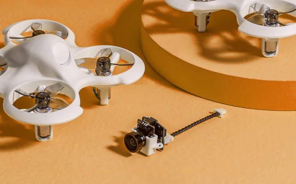 Kamera BETAFPV Cetus Lite Camera and VTX Module Mały czarny moduł kamery ze skręconym przewodem leży na pomarańczowym podłożu przed białym dronem z osłonami na śmigła