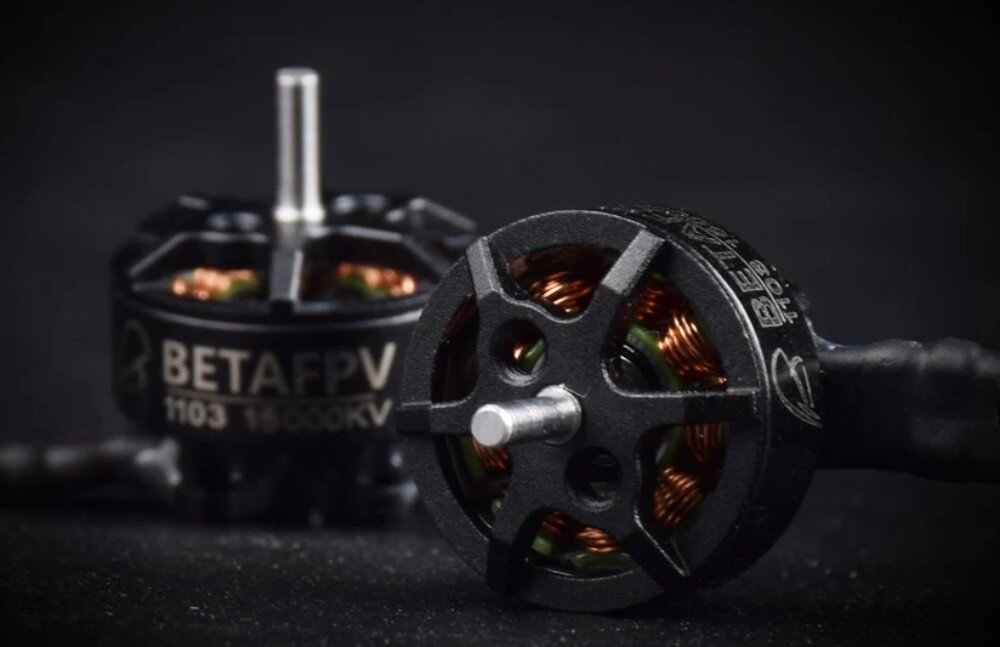 Silnik BETAFPV 1103 8500KV (4 szt.) Zbliżenie na dwa czarne silniki bezszczotkowe do drona leżące na ciemnym tle, ukazujące miedziane uzwojenie wewnątrz oraz srebrne wały wystające z górnej części obudowy