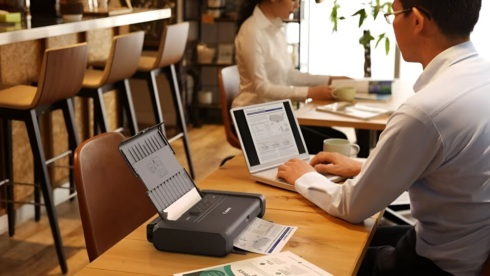 Drukarka CANON Maxify BX110 Druk w kolorze, Wi-Fi + Akumulator urządzenie na stole w kafejce mężczyzna siedzący z laptopem przy stole akumulator do 330 stron kabel USB-C gotowość do pracy podróż służbowa praca mobilna w terenie